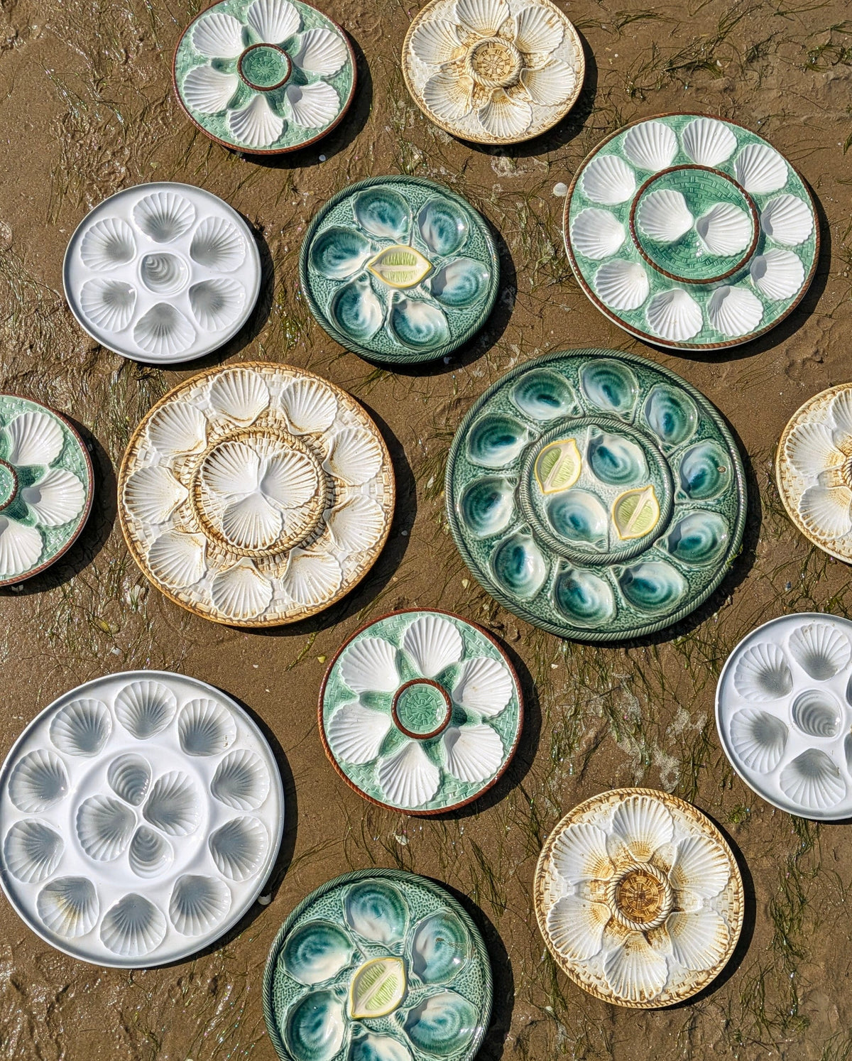 Collection d'assiettes à huîtres anciennes et vintage en faïence colorée, disposées sur le sable d'une plage.